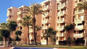 Una piscina rodeada por un edificio residencial de varios pisos con balcones, palmeras y un cielo despejado.