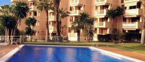 A swimming pool surrounded by a multi-story residential building with balconies, palm trees, and a clear sky.