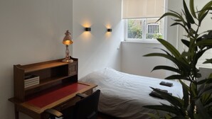 Quarto com escrivaninha de madeira, cadeira vermelha, cama branca e janela com persianas.