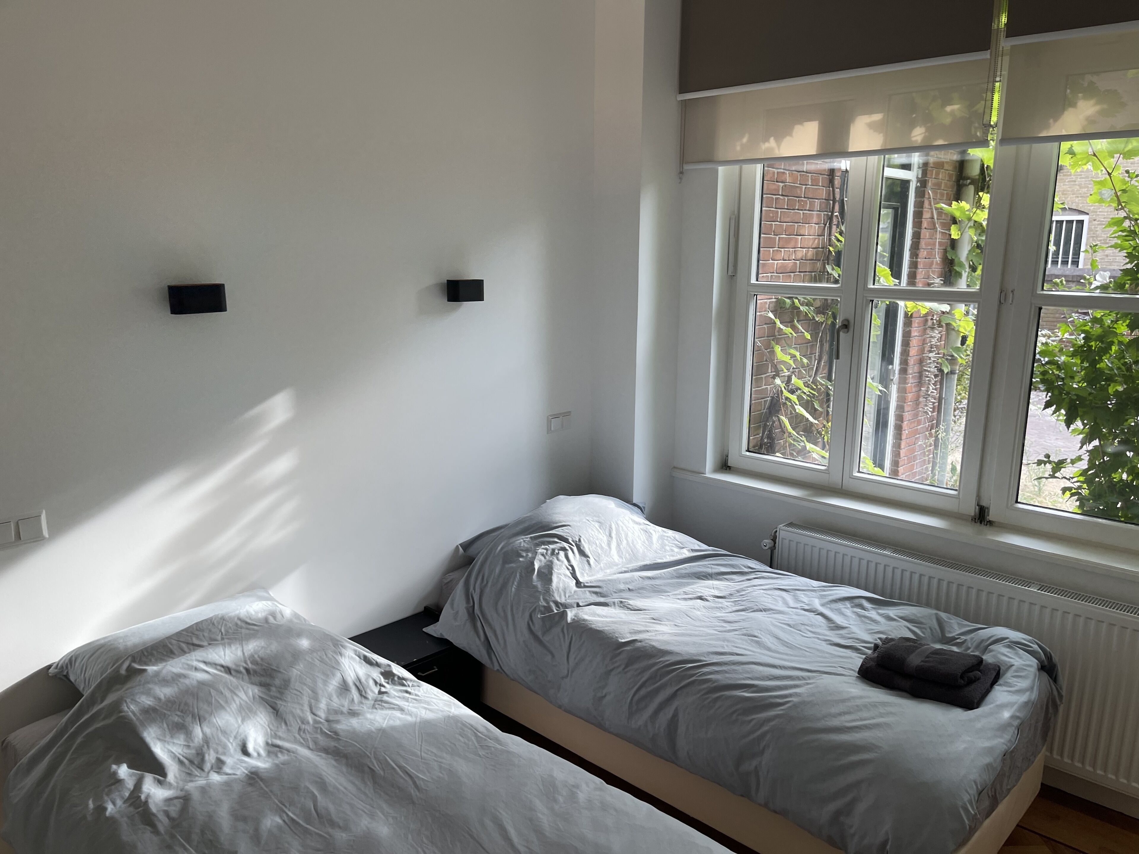 Een kleine, lichte kamer met twee eenpersoonsbedden, een radiator en een raam met uitzicht op een bakstenen gebouw en groen buiten.