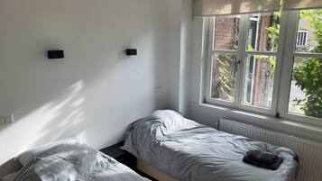 Een kleine, lichte kamer met twee eenpersoonsbedden, een radiator en een raam met uitzicht op een bakstenen gebouw en groen buiten.