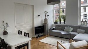 Uma sala de estar moderna com um sofá cinzento, uma mesa de jantar branca com cadeiras, uma televisão e uma janela grande com vista para a cidade.