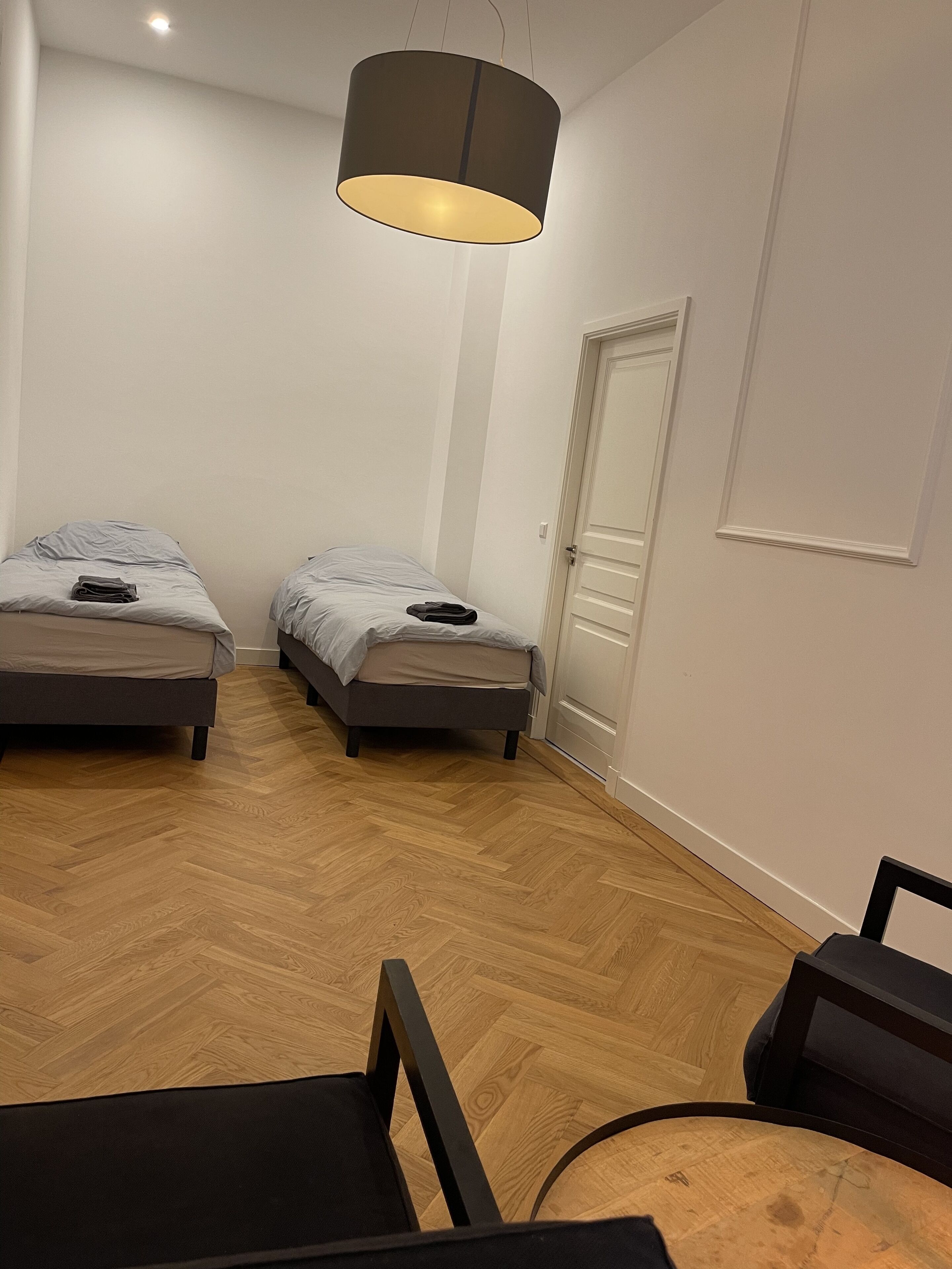 A room with two beds, a wooden floor, a door, and a hanging light fixture.