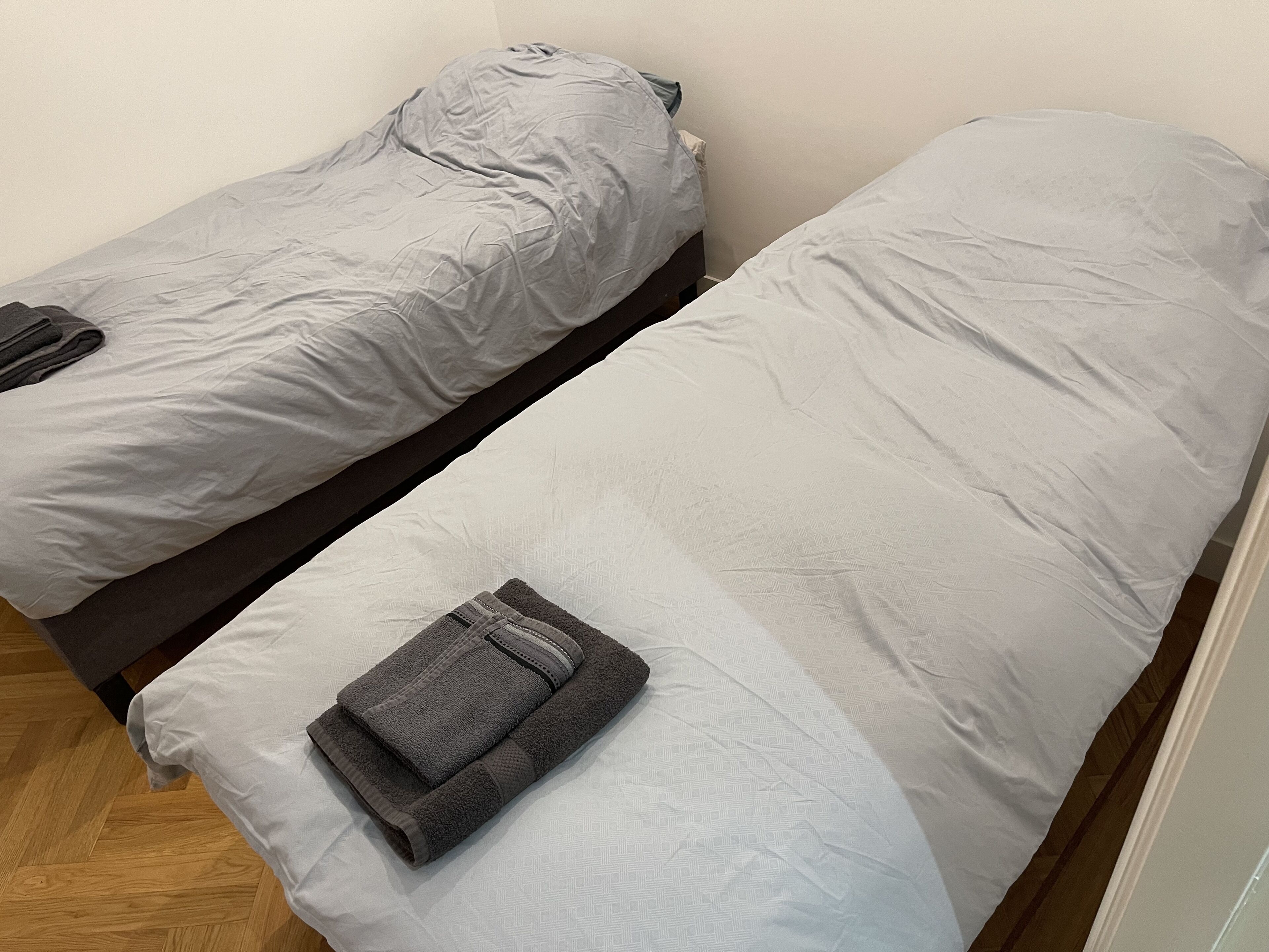 Dos camas individuales con ropa de cama gris, una toalla gris doblada sobre una cama, y piso de madera.