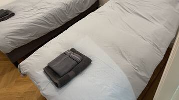 Dos camas individuales con ropa de cama gris, una toalla gris doblada sobre una cama, y piso de madera.