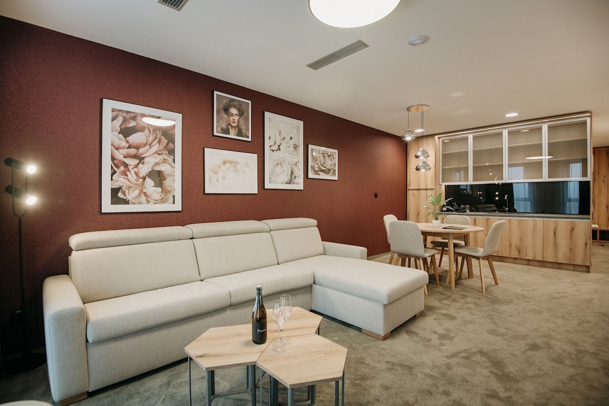 Una sala de estar moderna con un sofá seccional color beige, una pequeña mesa de madera con una botella y vasos, y un comedor con una mesa y sillas de madera.