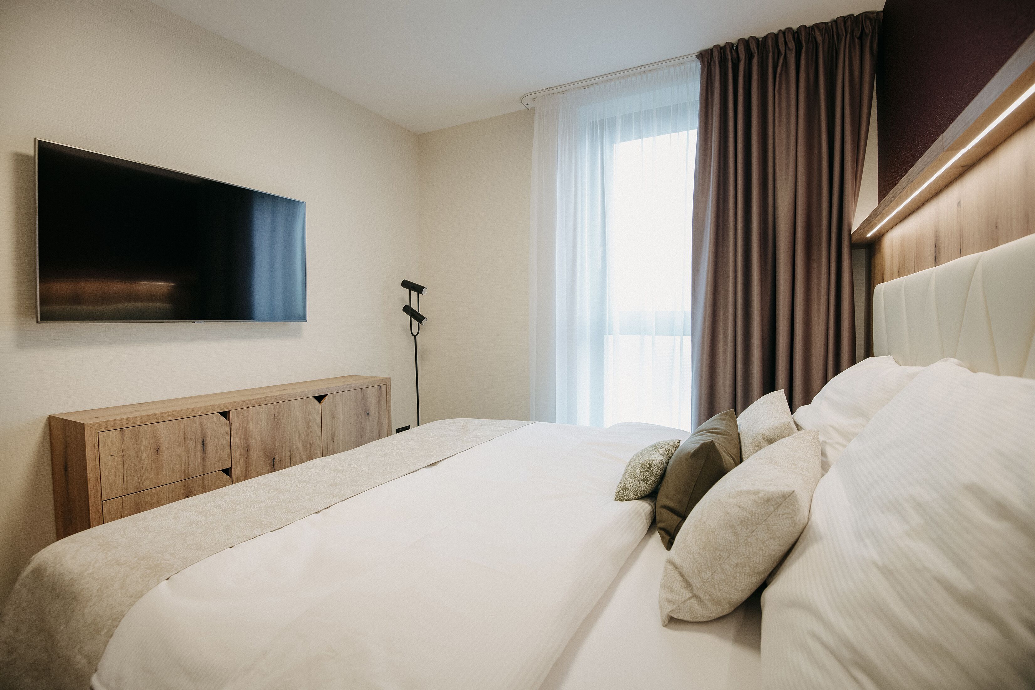 A modern hotel room with a large bed, a flat-screen TV mounted on the wall, and a window with curtains.