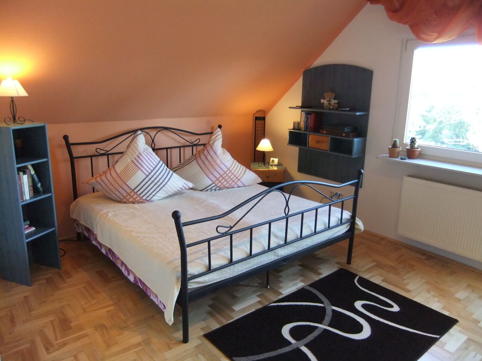 A bedroom with a bed, bedside lamps, a bookshelf, a small shelf with books and a teddy bear, a window with a view, and a radiator.