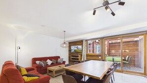 A modern living room with a red sofa, a wooden dining table, and a large window with a view of the outdoors. - Luxurious 2-Bedroom Apartment in New Residence, Sleeps 6 – Heart of Les Gets (Les Gets)