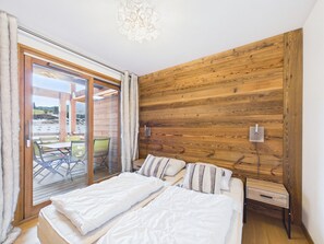 Een slaapkamer met een groot bed, een houten wand, een schuifdeur van glas naar een buitenruimte en een klein nachtkastje.