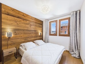 A bedroom with a wooden wall, a bed with white bedding, a bedside table, and two windows with curtains.