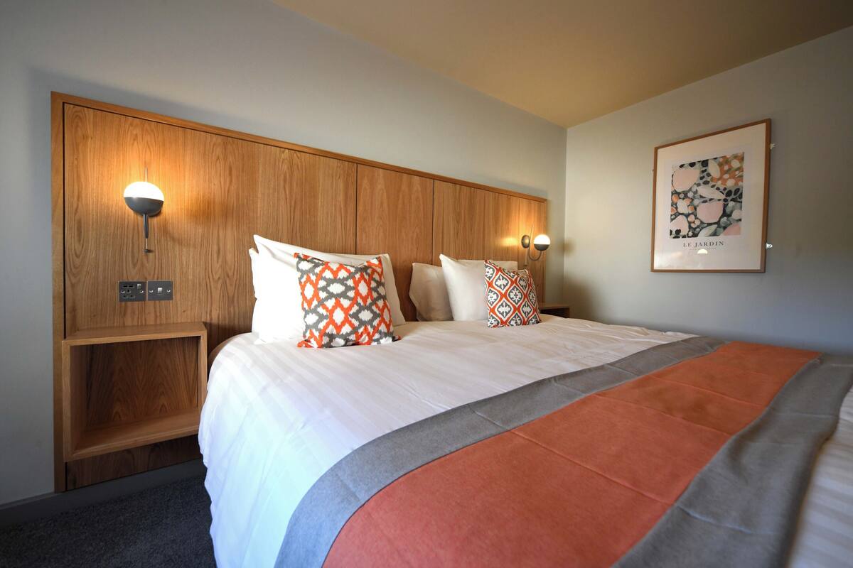 A hotel room with a large bed, two bedside tables, a wooden headboard, and two wall-mounted lamps.