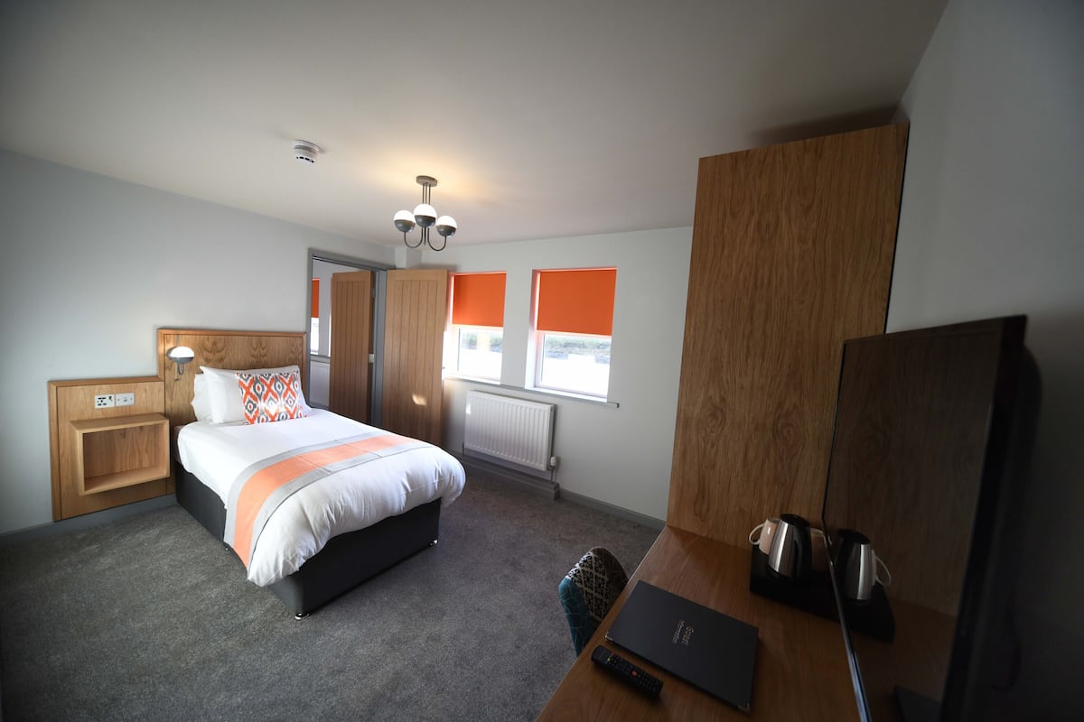 A modern hotel room with a bed, a desk, a chair, a television, and a window with orange blinds.