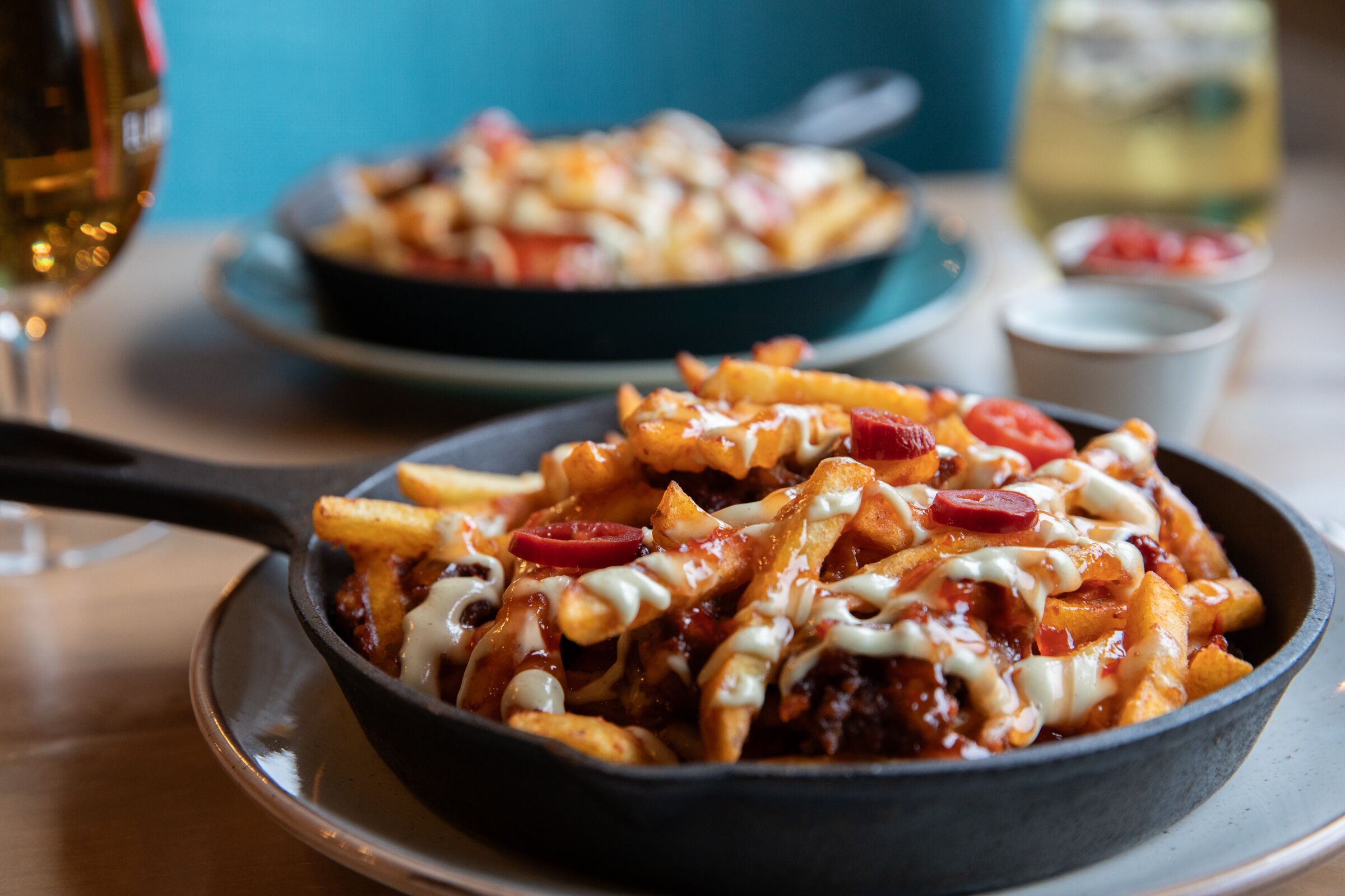 A skillet of loaded fries with toppings, a glass of wine, and a bowl of salad on a table.