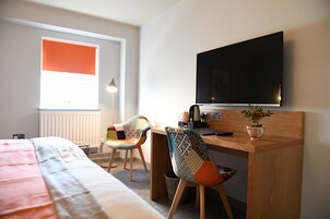Una habitación de hotel con cama, escritorio con televisor, silla, lámpara y ventana con persianas.