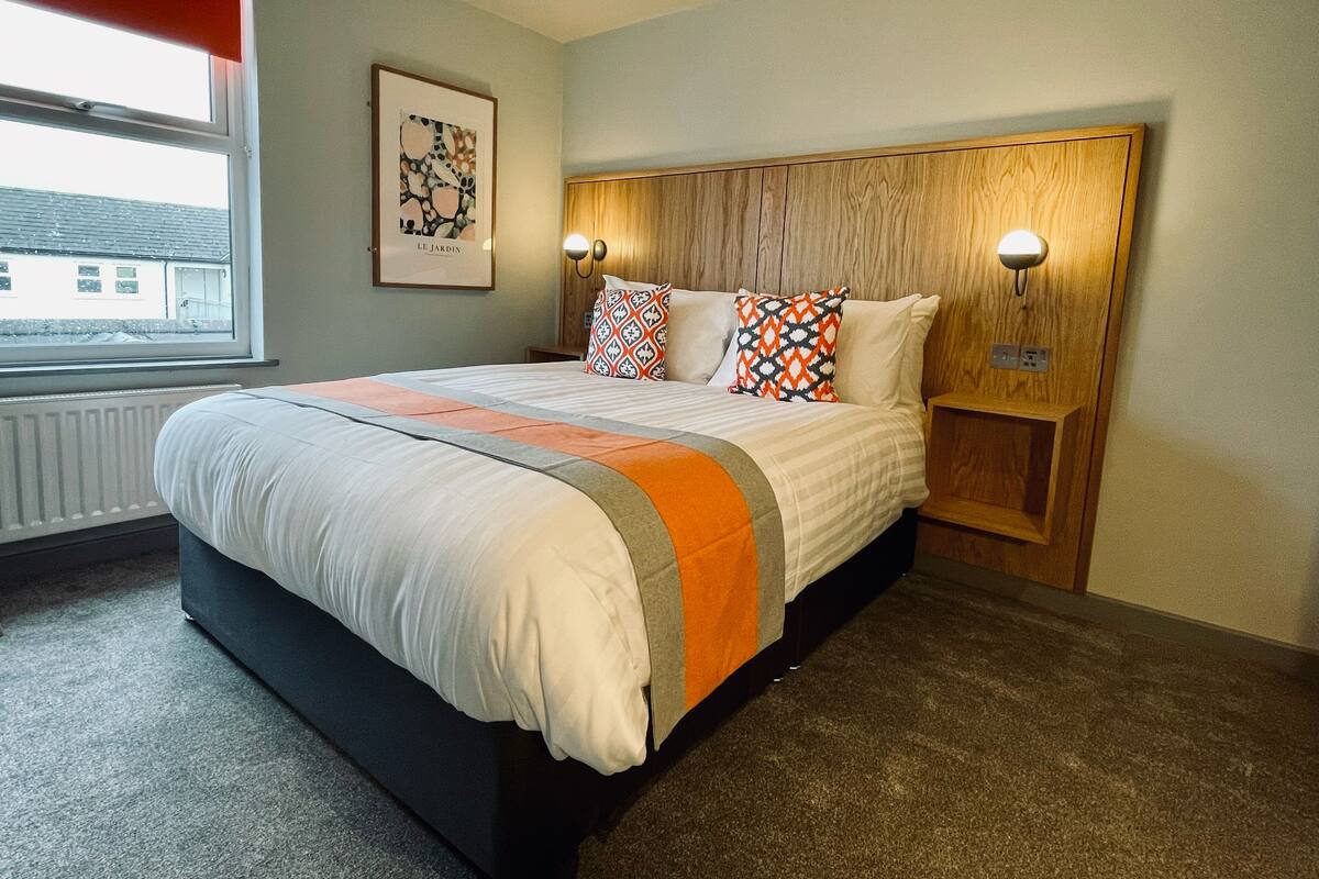 A neatly made bed with a patterned blanket and pillows, a wooden headboard with wall-mounted lights, a radiator under the window, and a framed picture on the wall.