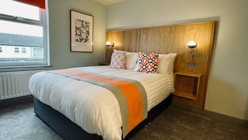 Una cama bien hecha con una colcha y almohadas estampadas, cabecera de madera con luces fijadas a la pared, radiador debajo de la ventana y un cuadro enmarcado en la pared.