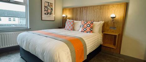 A neatly made bed with a patterned blanket and pillows, a wooden headboard with wall-mounted lights, a radiator under the window, and a framed picture on the wall.