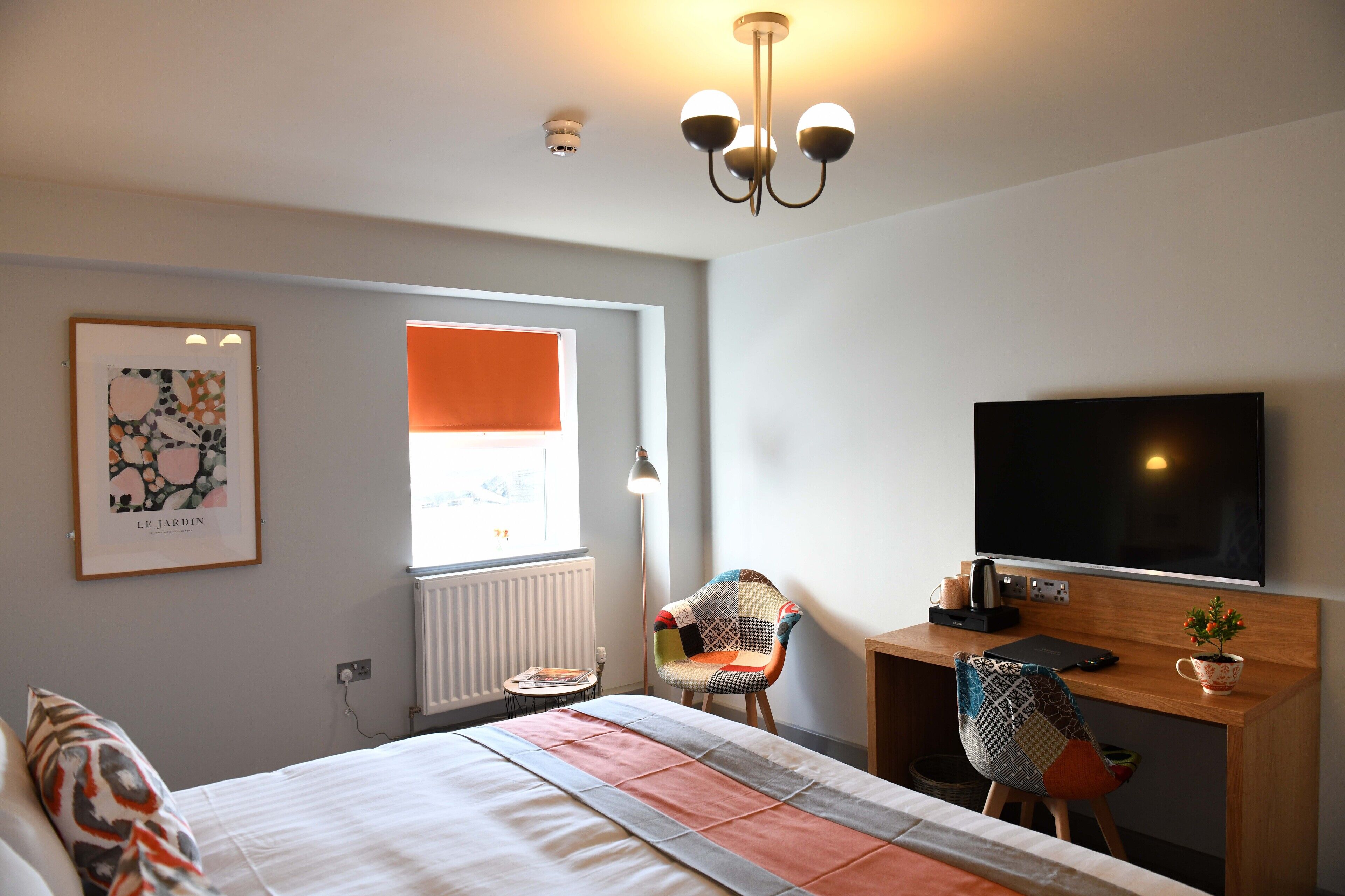 A hotel room with a bed, a desk with a TV, a chair, a window with orange blinds, and a radiator.