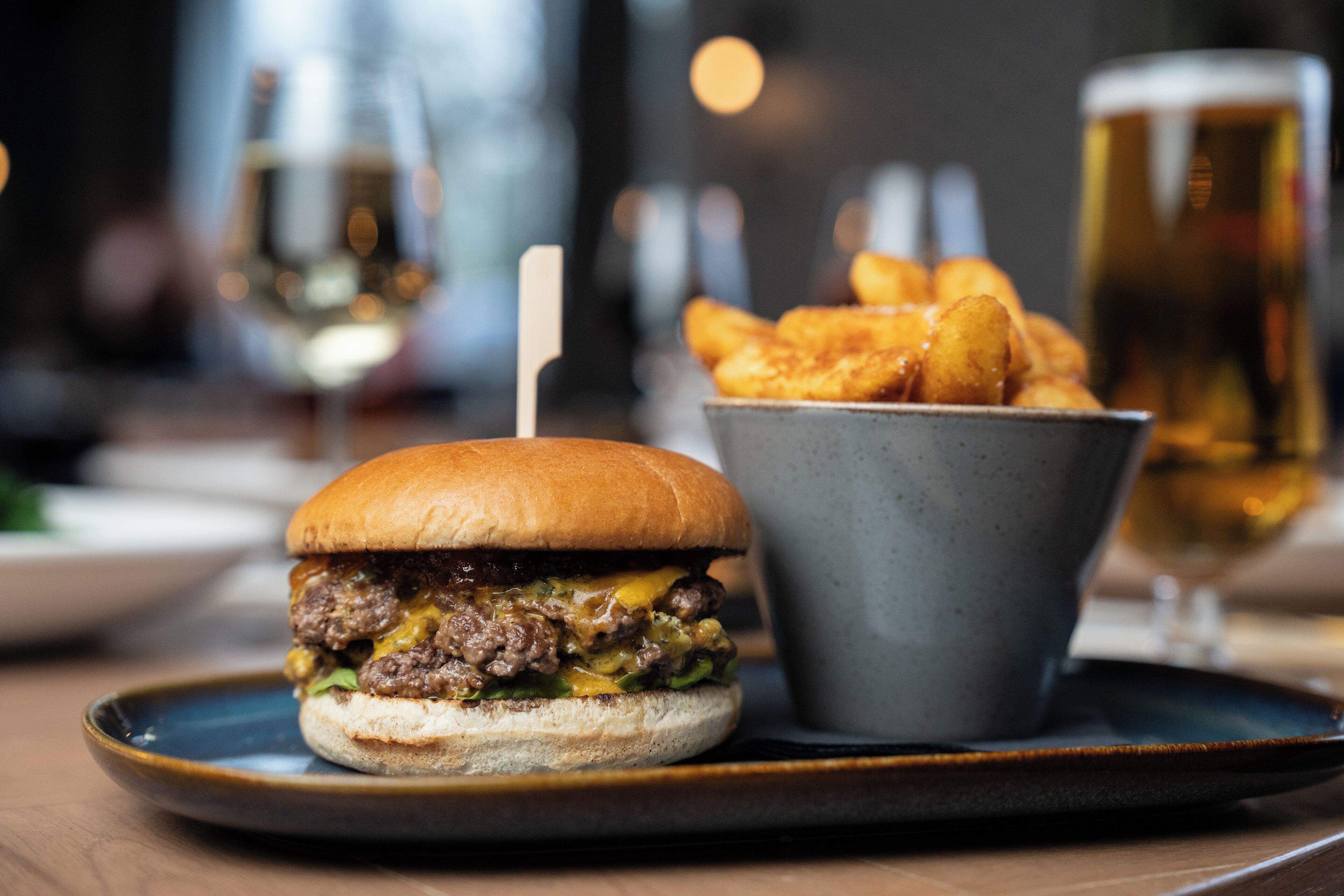 Una hamburguesa con papas fritas en un plato, con una jarra de cerveza al fondo.