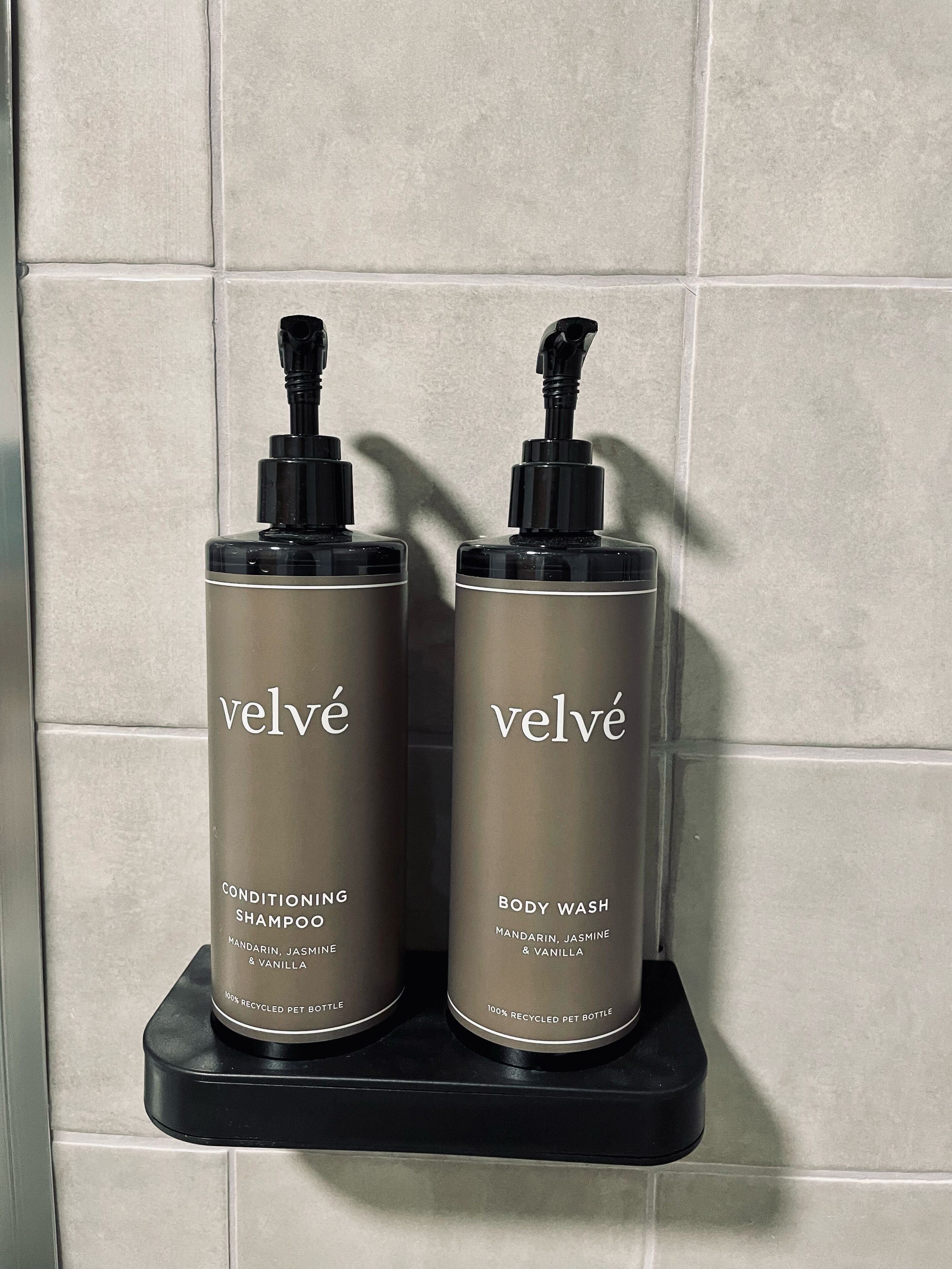 Deux bouteilles de shampoing et de gel douche Velvé, posées sur une étagère noire devant un mur carrelé.
