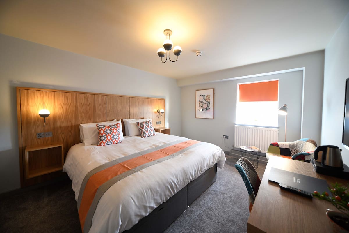 A modern hotel room with a large bed, a desk with a laptop, a coffee maker, and a view of the city.