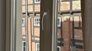 A window with a view of a brick building, a metal pole, and a glass vase with items on a windowsill. - Welcome to the harbor district - Emma (Wismar)