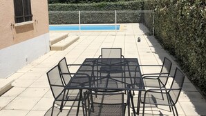 Un patio con mesa y sillas, una piscina y un área cercada con árboles y arbustos.