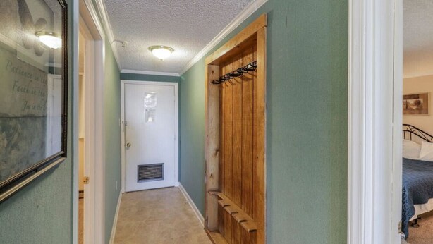 Een smalle gang met een deur aan het einde, een houten kast met kapstok, en een bed in de aangrenzende kamer.