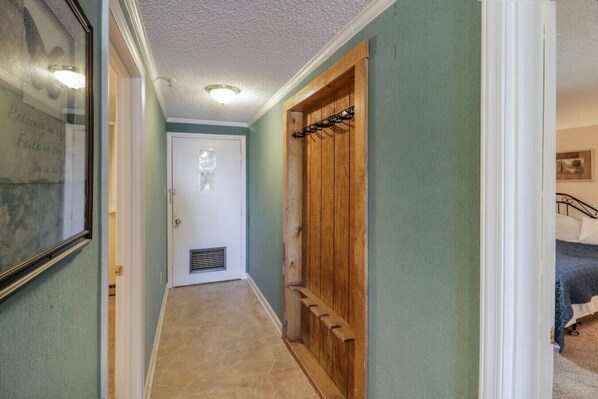 A narrow hallway with a door at the end, a wooden closet with a coat rack, and a bed in the adjacent room.