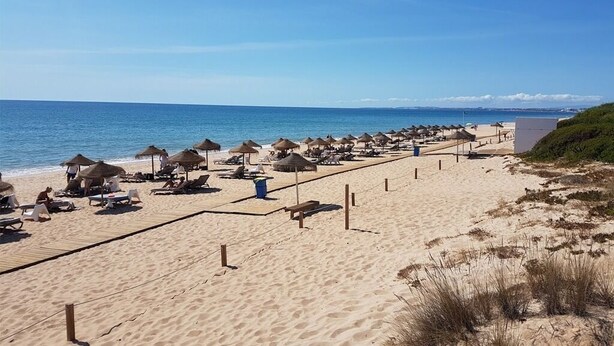 Ein Strand mit Liegestühlen und Sonnenschirmen, ein Holzsteg und freie Sicht auf den Ozean.