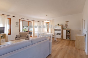 A modern living room with a sofa, dining table, and chairs. There is a kitchen area with cabinets and a refrigerator.