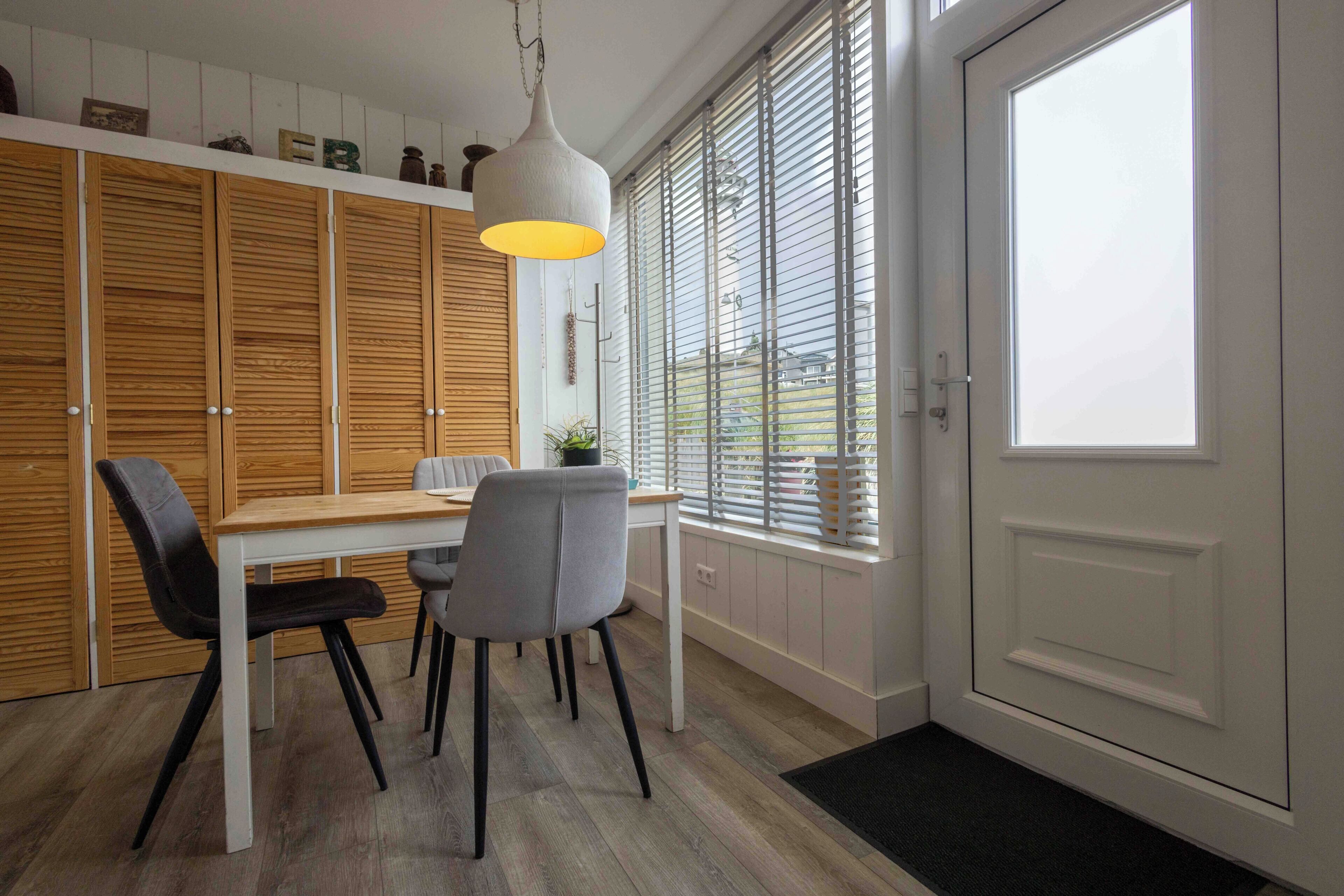 A dining area with a wooden table, two chairs, a pendant light, and a view through a window with blinds.