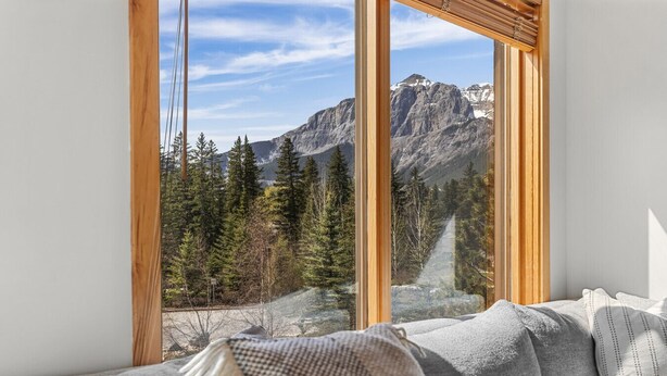 A room with a large window offering a mountain view, a gray sofa with a throw blanket, and wooden window frames.