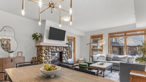 A modern living room with a stone fireplace, a dining area with a wooden table, and a large window offering a view of the outside.