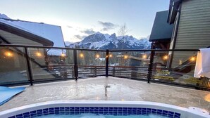 A hot tub with a view of snow-capped mountains, a glass enclosure, and a wooden deck.