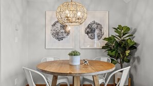 Een eetgedeelte met een ronde houten tafel, witte stoelen, een hanglamp en twee kunstwerken in lijsten aan de muur.