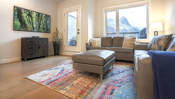 Un salon avec un canapé, un pouf et un tapis coloré. Il y a une grande fenêtre donnant sur les montagnes, une porte et une plante.