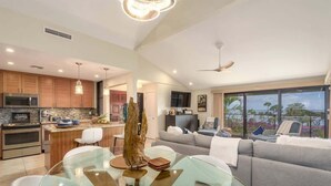 Una cocina moderna con una isla central, un comedor con mesa de vidrio y una sala de estar con sofá y ventanas amplias.