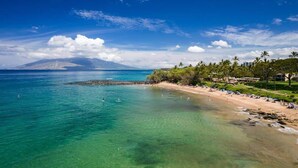 Una playa con aguas turquesas cristalinas, una orilla arenosa y montañas con palmeras al fondo.