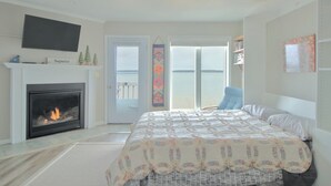 Quarto com lareira, cama com colcha floral, uma cadeira azul e vista para o mar através de janelas amplas.