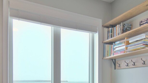 Quarto com uma grande janela com vista para a água, uma poltrona estofada azul e uma estante com livros e ganchos na parede.