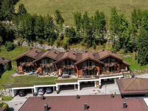 A multi-story wooden lodge with balconies, a covered parking area, and a view of a green hillside.