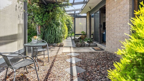 A covered outdoor area with a patio table and chairs, surrounded by greenery and a brick wall.