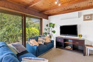 A living room with a blue sofa, a flat-screen TV, a wooden ceiling, and a large window with blinds.