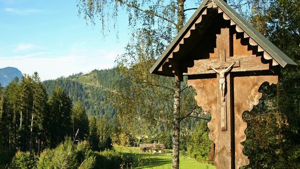 Une structure en bois en forme de crucifix, surmontée d’un toit triangulaire, se dressant sur un fond de paysage montagneux et de ciel dégagé.