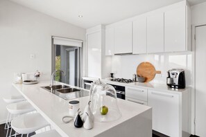 A modern kitchen with white cabinetry, a central island, a stove, and a coffee maker. - Summerhills House (Lorne)