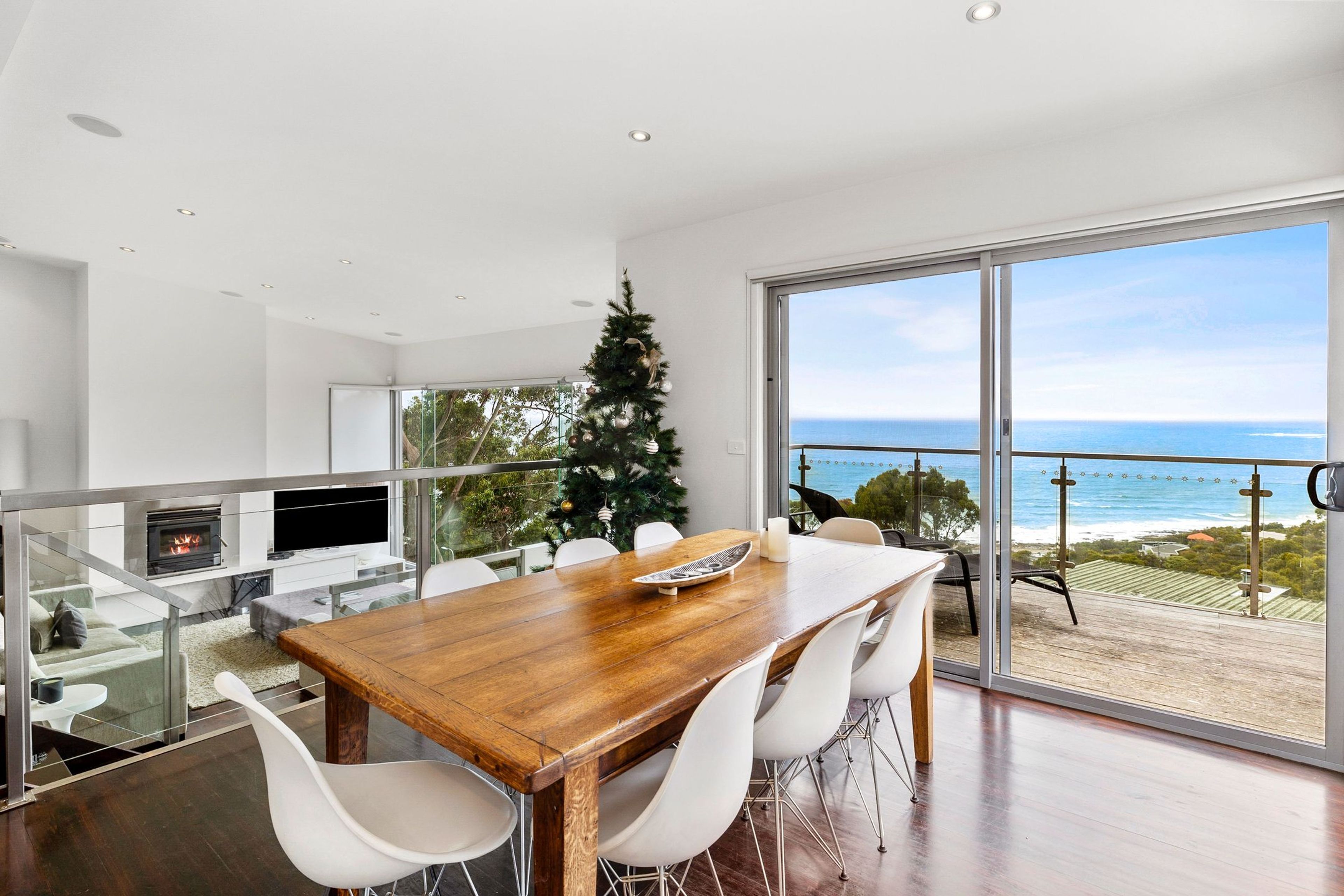 A modern dining area with a wooden table and white chairs, a fireplace, and a large window overlooking the ocean.