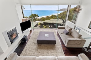A modern living room with a large TV, comfortable seating, a fireplace, and a view of the ocean. - Summerhills House (Lorne)