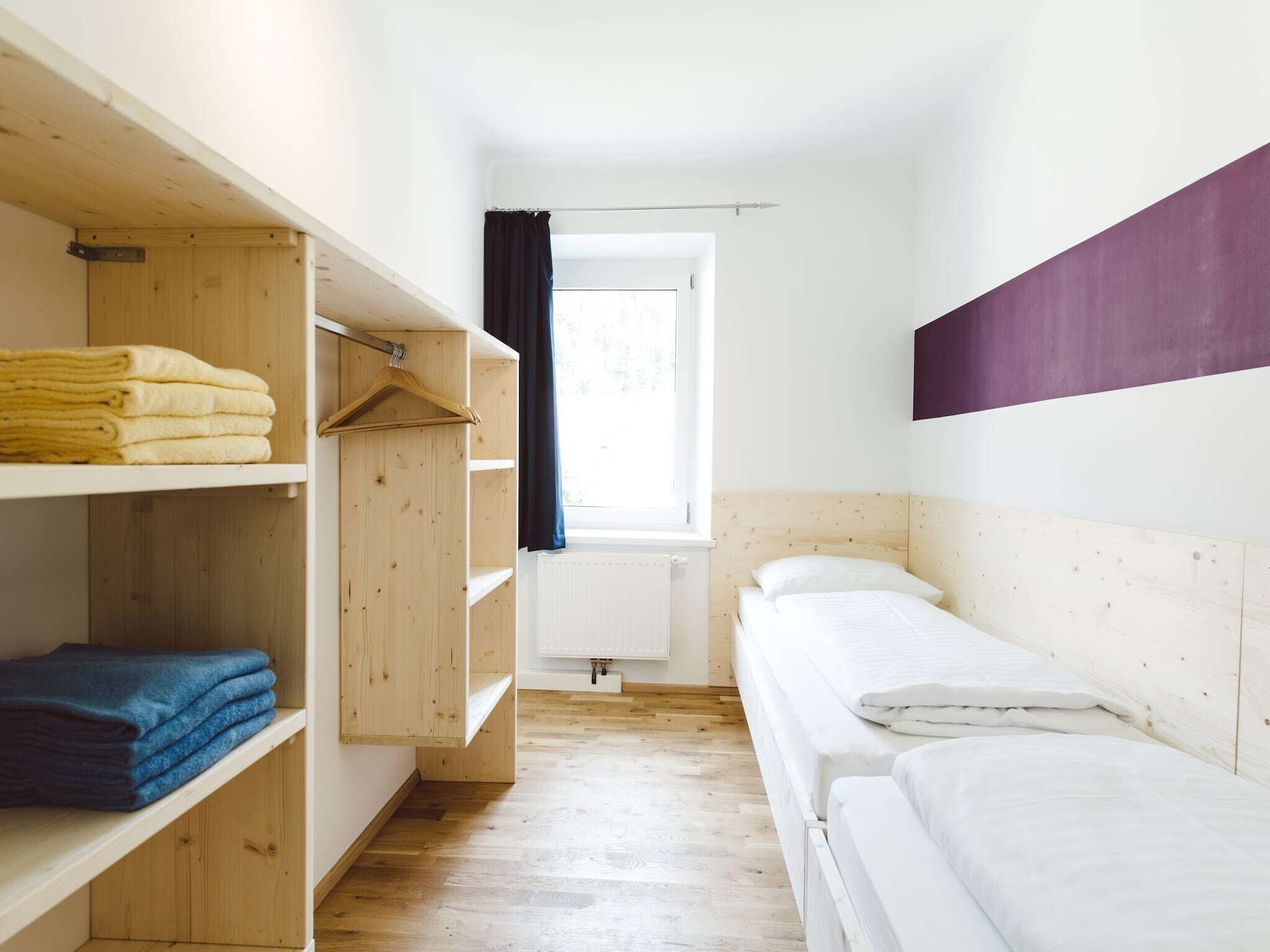 A small, well-lit room with a single bed, wooden shelves, and a window with curtains.